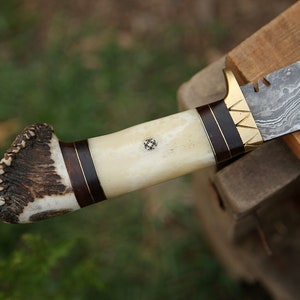 May include: A hunting knife with a damascus steel blade and a handle made of antler and wood. The handle has a brass accent and a small decorative inlay.