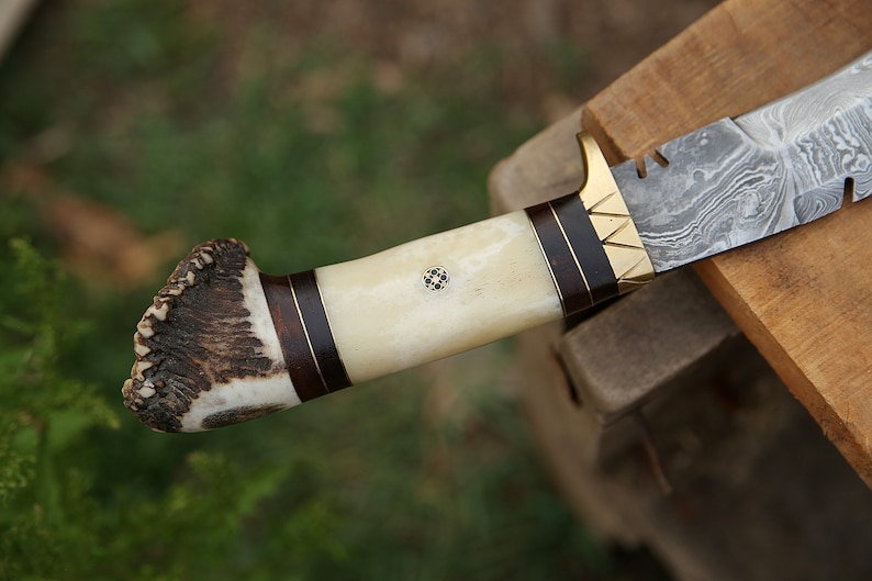 May include: A hunting knife with a damascus steel blade and a handle made of antler and wood. The handle has a brass accent and a small decorative inlay.