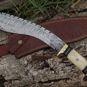 May include: A curved, silver-colored knife with a brown leather sheath. The knife has a patterned blade and a white handle with a brown antler tip. The sheath has a brown leather strap.