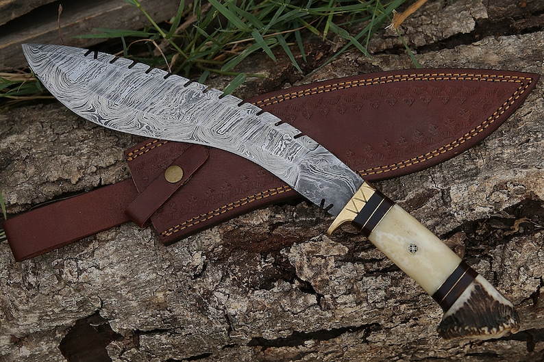 May include: A curved, silver-colored knife with a brown leather sheath. The knife has a patterned blade and a white handle with a brown antler tip. The sheath has a brown leather strap.