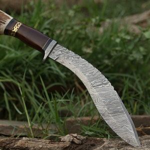 May include: A curved, silver-colored knife with a brown wooden handle and a stag antler pommel. The knife has a Damascus steel blade with a wavy pattern.