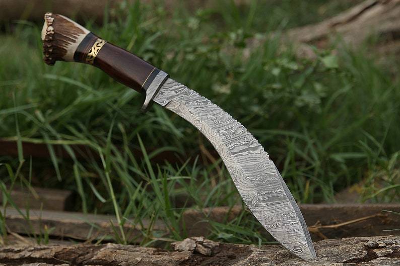 May include: A curved, silver-colored knife with a brown wooden handle and a stag antler pommel. The knife has a Damascus steel blade with a wavy pattern.