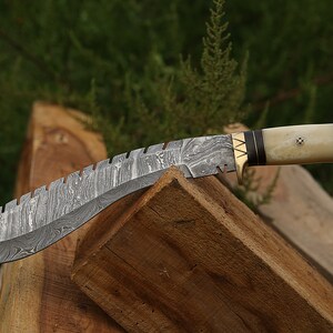 May include: A curved, silver-colored knife with a patterned blade and a brown and white handle. The handle is made of wood and antler. The knife is resting on a piece of wood.