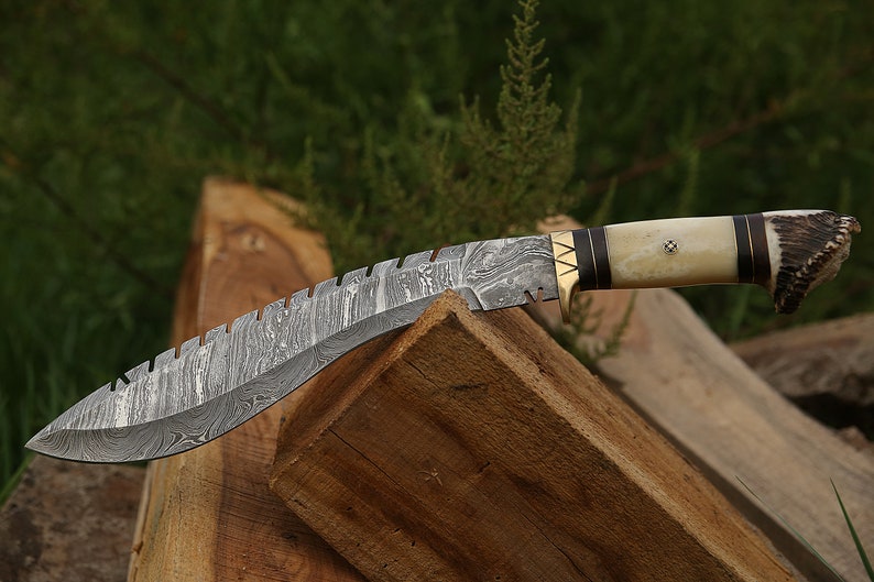 May include: A curved, silver-colored knife with a patterned blade and a brown and white handle. The handle is made of wood and antler. The knife is resting on a piece of wood.