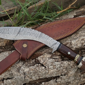 May include: A curved, silver-colored knife with a brown wooden handle and a leather sheath. The knife has a decorative pattern on the blade and a gold accent on the handle.