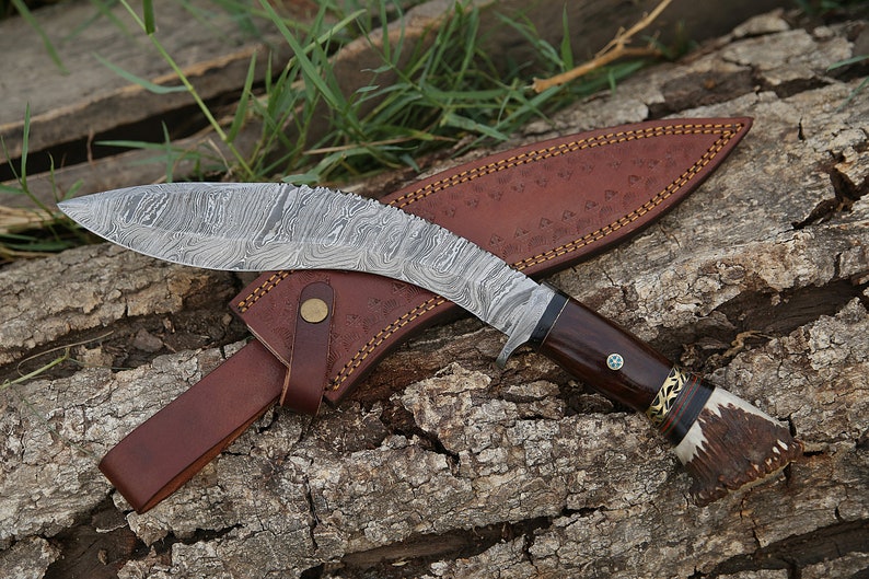 May include: A curved, silver-colored knife with a brown wooden handle and a leather sheath. The knife has a decorative pattern on the blade and a gold accent on the handle.