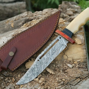 May include: A hunting knife with a brown leather sheath and a white bone handle. The blade is made of Damascus steel and has a serrated edge.