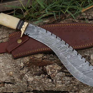 May include: A curved, silver-colored knife with a patterned blade and a brown leather sheath. The handle is made of bone and antler with a brass accent.