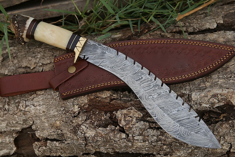 May include: A curved, silver-colored knife with a patterned blade and a brown leather sheath. The handle is made of bone and antler with a brass accent.