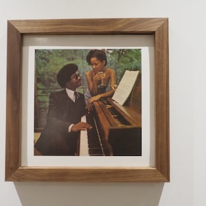Puede incluir: Una fotografía enmarcada de una pareja, un hombre tocando un piano y una mujer sentada a su lado, sosteniendo una copa de vino. La foto está ambientada en un jardín verde exuberante.