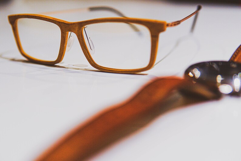 K&ouml;nnte beinhalten: Eine Brille mit braunem Holzrahmen und klaren Gl&auml;sern. Die Brille liegt auf einer wei&szlig;en Oberfl&auml;che mit einem braunen Lederband im Vordergrund.