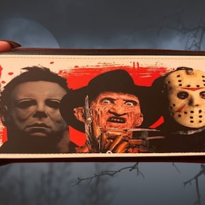 May include: A rectangular wallet featuring Michael Myers, Freddy Krueger, and Jason Voorhees. The design has a white background with red splatter effects. The wallet has a black border and a brown interior.