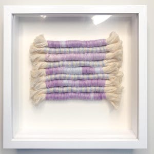 May include: A fiber art piece displayed in a square white shadow box frame. The artwork consists of several horizontal rows of cream-colored cord wrapped with variegated yarn in shades of light purple, lavender, and soft blue, with frayed ends.