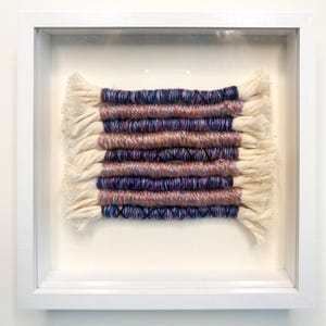 May include: A square white shadow box frame containing a woven fiber art piece. The textile features horizontal rows of yarn wrapped around thick cream-colored cotton cords, alternating between deep navy blue and variegated peach and purple tones.