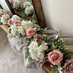 May include: A floral garland featuring white hydrangeas, dusty rose roses, and green foliage. The arrangement is displayed on a wooden surface, with a neutral-toned blanket underneath. The garland is reflected in a wooden-framed mirror.