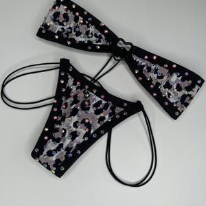 May include: A two-piece bikini set featuring a black, white, and gray leopard print pattern. Both the bandeau top and the bottom are embellished with numerous iridescent rhinestones and finished with black trim and thin black side straps.