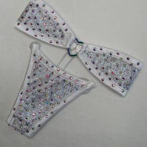 May include: White bikini set with glitter and iridescent rhinestones. The top features a halter design with a connecting iridescent ring. The bottoms are a classic cut. The edges of the bikini are lined with rhinestones.