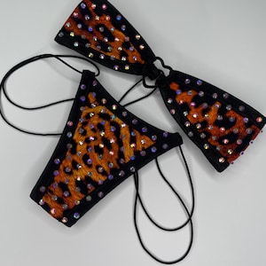 May include: A two-piece bikini set featuring a black and orange leopard print pattern. Both the top and the bottom are embellished with numerous iridescent rhinestones and finished with black trim and thin black string ties for adjustment.