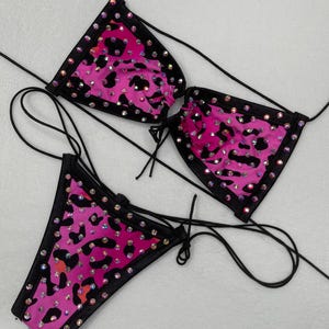 May include: A three-piece bikini set featuring a pink leopard print fabric with black trim. The bikini top and bottom are adorned with iridescent rhinestones. Black string ties are used for the top and bottom.
