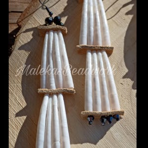 May include: Handmade white beaded earrings with a leather accent and black bead details. The earrings are displayed on a wooden surface. The text "Malekas Beadwork" is visible on the wood.
