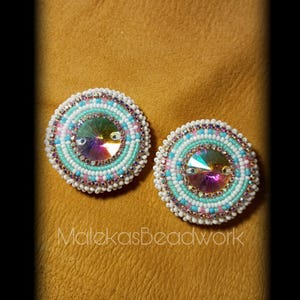 May include: Two round beaded earrings with a central iridescent crystal. The earrings feature a pattern of white, light blue, and pink beads, surrounded by a border of smaller beads. The text "MalekasBeadwork" is at the bottom.