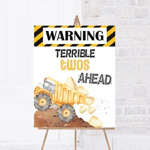 Könnte beinhalten: Ein gelber Bauwagen mit einem Warnschild, auf dem "Achtung Schreckliche Zweier vor uns" steht. Das Schild ist auf weißem Hintergrund mit schwarzen und gelben Streifen.