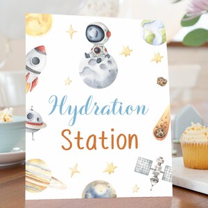 May include: A white sign with watercolor illustrations of planets, rockets, and an astronaut on a moon. The sign reads "Hydration Station" in blue and orange script. The sign is on a wooden table with cupcakes.