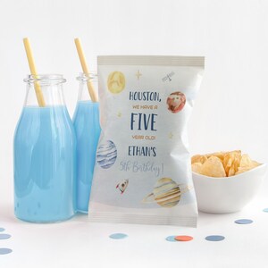 Puede incluir: Una bolsa de patatas fritas blanca con un diseño espacial azul y amarillo. La bolsa tiene el texto "Houston, we have a FIVE year old! ETHAN'S 5th Birthday!" impreso en ella. La bolsa está sentada sobre una superficie blanca con confeti azul y naranja.