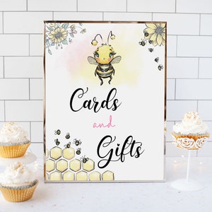 Puede incluir: Un cartel blanco con marco plateado que presenta una abeja de dibujos animados con un lazo rosa y el texto "Cards and Gifts" en letra cursiva negra. El cartel está sobre una superficie de azulejos blancos con dos cupcakes en un soporte dorado.