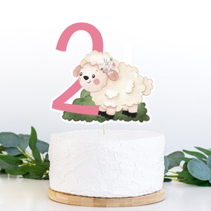May include: A pink number two cake topper with a white lamb with a floral crown. The lamb is standing on a green patch of grass.