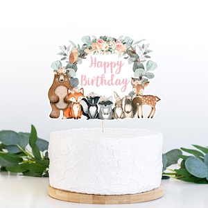 Op de afbeelding: Een taarttopper met een bosdierenthema met een roze bloemenkrans en de tekst "Happy Birthday". De topper bevat een beer, een vos, een wasbeer, een konijn en een hert.