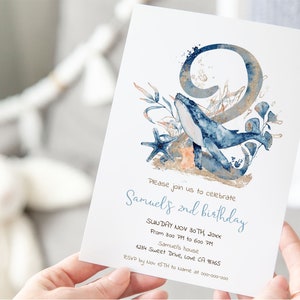 Op de afbeelding: Een blauwe en gouden uitnodigingskaart met een aquarel-illustratie van een walvis die door zeewier zwemt en een groot nummer 2. De tekst op de kaart luidt "We nodigen u uit om de 2e verjaardag van Samuels te vieren, zondag 30 november 20xx, van 15:00 tot 18:00 uur, bij Samuels, 1234 Sweet Drive, Love CA 98765, RSVP v&oacute;&oacute;r 15 november aan Name op 000-000-000".