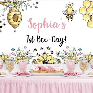Puede incluir: Un telón de fondo rosa y blanco con un diseño de abeja y panal. El texto "Sophia's 1st Bee-Day!" está escrito en negro. Una mesa con un mantel rosa está preparada con tarros de dulces, cupcakes y galletas.