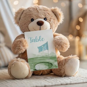 Pode incluir: Um urso de peluche castanho segurando um cartão com o texto "Mesa 1" e uma ilustração de aquarela de um campo de golfe com uma bandeira.