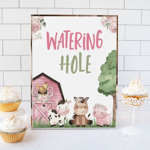 May include: A pink and green watercolor sign with the text "Watering Hole" and illustrations of a red barn, a cow, a horse, and a pig. The sign is on a white tiled surface with cupcakes on a stand.