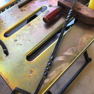 May include: A twisted metal rod with a hook on one end and a pointed end on the other. The rod is lying on a metal workbench with a wooden clamp and a red handle.