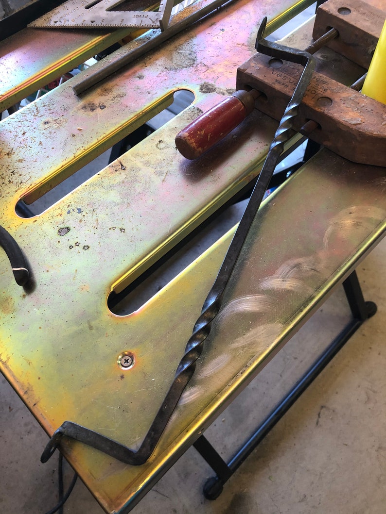 May include: A twisted metal rod with a hook on one end and a pointed end on the other. The rod is lying on a metal workbench with a wooden clamp and a red handle.