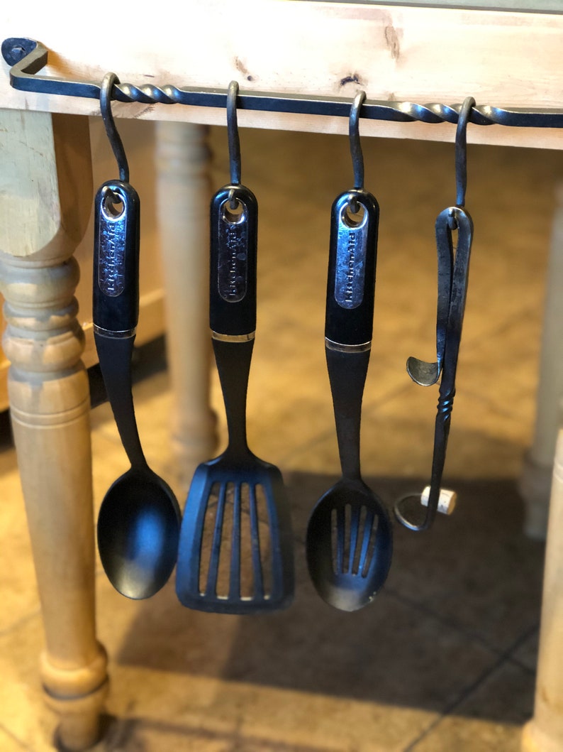 May include: Four black kitchen utensils hanging on a metal rack with a twisted design. The utensils include a slotted spoon, a spatula, a spoon, and a metal tool.