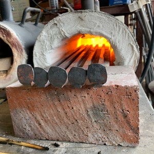 May include: Close-up of a metal forge with a brick base. The forge is lit and glowing orange. Several metal rods with rounded ends are placed in the forge.