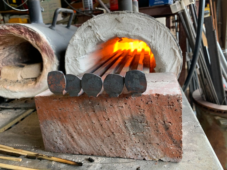 May include: Close-up of a metal forge with a brick base. The forge is lit and glowing orange. Several metal rods with rounded ends are placed in the forge.