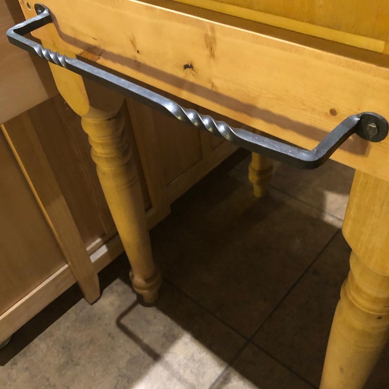 May include: A black metal towel rack with a twisted design is mounted to a wooden countertop. The rack is attached to the countertop with two screws.