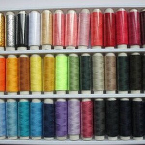 K&ouml;nnte beinhalten: Ein Set mit 36 Garnrollen in verschiedenen Farben, darunter Gold, Silber, Rot, Gr&uuml;n, Blau und Lila. Die Rollen sind in einem Plastikkoffer mit einem durchsichtigen Deckel angeordnet.