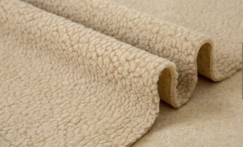 May include: Close-up of a beige faux fur fabric with a soft, fuzzy texture. The fabric is folded over itself, revealing a smooth, brown underside.