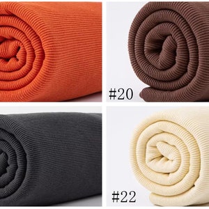 Op de afbeelding: Vier rollen geribbelde stof in verschillende kleuren: oranje, bruin, zwart en cr&egrave;me. De stof is opgerold en de uiteinden zijn zichtbaar. De nummers #19, #20, #21 en #22 zijn onder elke rol afgedrukt.