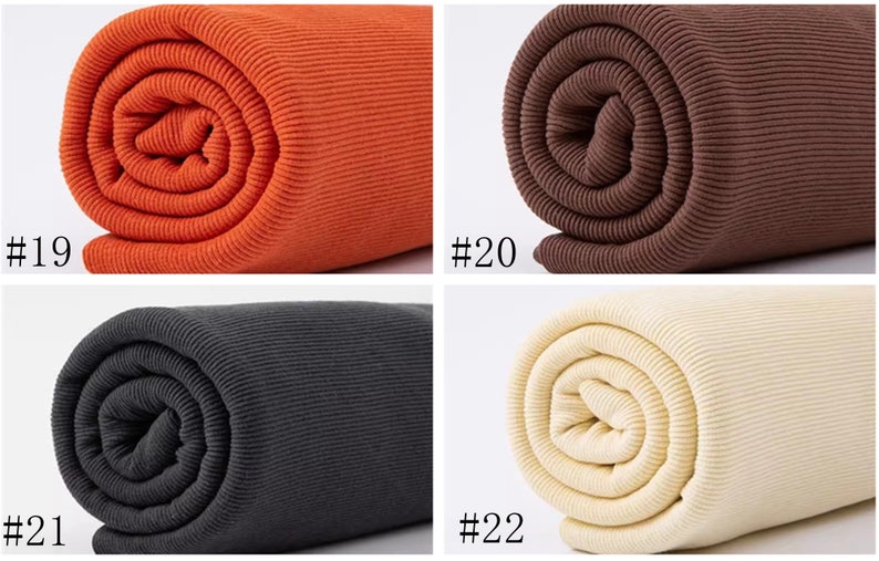Op de afbeelding: Vier rollen geribbelde stof in verschillende kleuren: oranje, bruin, zwart en cr&egrave;me. De stof is opgerold en de uiteinden zijn zichtbaar. De nummers #19, #20, #21 en #22 zijn onder elke rol afgedrukt.