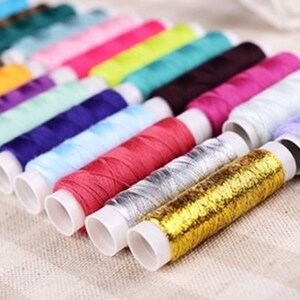 K&ouml;nnte beinhalten: Eine Sammlung von bunten Garnrollen, darunter Gold, Silber, Rot, Lila, Blau, Gr&uuml;n und Gelb. Die Rollen sind in einer Reihe auf einer beigefarbenen Stoffoberfl&auml;che angeordnet.