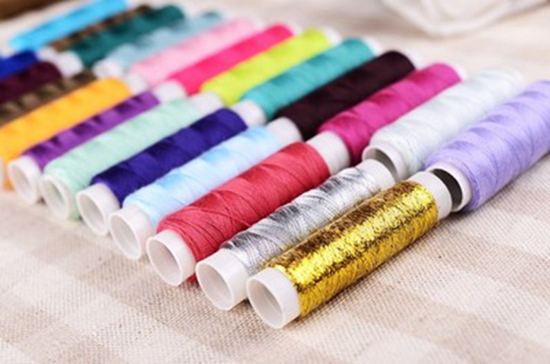 K&ouml;nnte beinhalten: Eine Sammlung von bunten Garnrollen, darunter Gold, Silber, Rot, Lila, Blau, Gr&uuml;n und Gelb. Die Rollen sind in einer Reihe auf einer beigefarbenen Stoffoberfl&auml;che angeordnet.