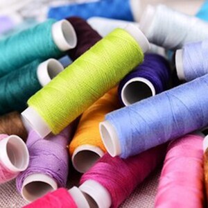 K&ouml;nnte beinhalten: Eine Sammlung bunter Garnrollen, darunter rosa, lila, blau, gr&uuml;n, gelb, orange, braun und gold. Die Rollen sind in einem Stapel angeordnet, wobei sich einige &uuml;berlappen.