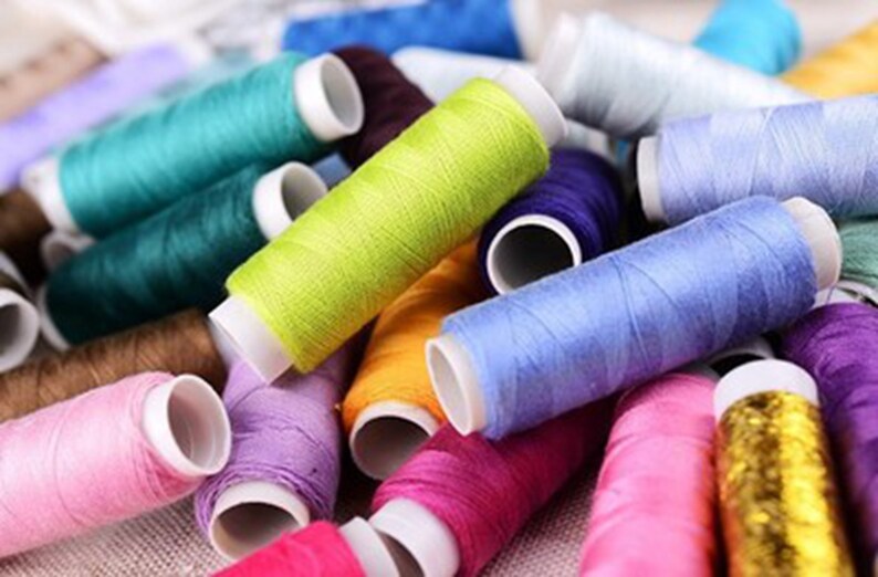 K&ouml;nnte beinhalten: Eine Sammlung bunter Garnrollen, darunter rosa, lila, blau, gr&uuml;n, gelb, orange, braun und gold. Die Rollen sind in einem Stapel angeordnet, wobei sich einige &uuml;berlappen.