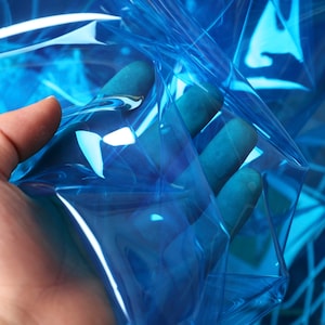 Może przedstawiać: Arkusz przezroczystego, niebieskiego plastiku z lekkim, tęczowym połyskiem. Plastik jest złożony i trzymany w dłoni.